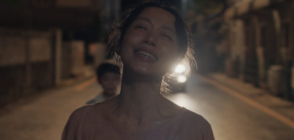 Smiling woman standing on a dimly lit street at night as car headlights glow behind her