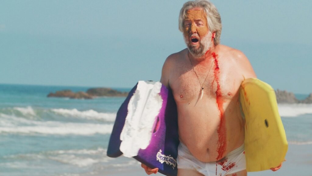 Older man with a bleeding chest wound shouts while clutching a surfboard on a beach