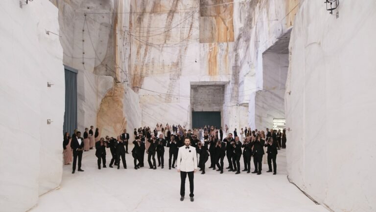 Man in white jacket stands before a crowd in a vast white marble quarry