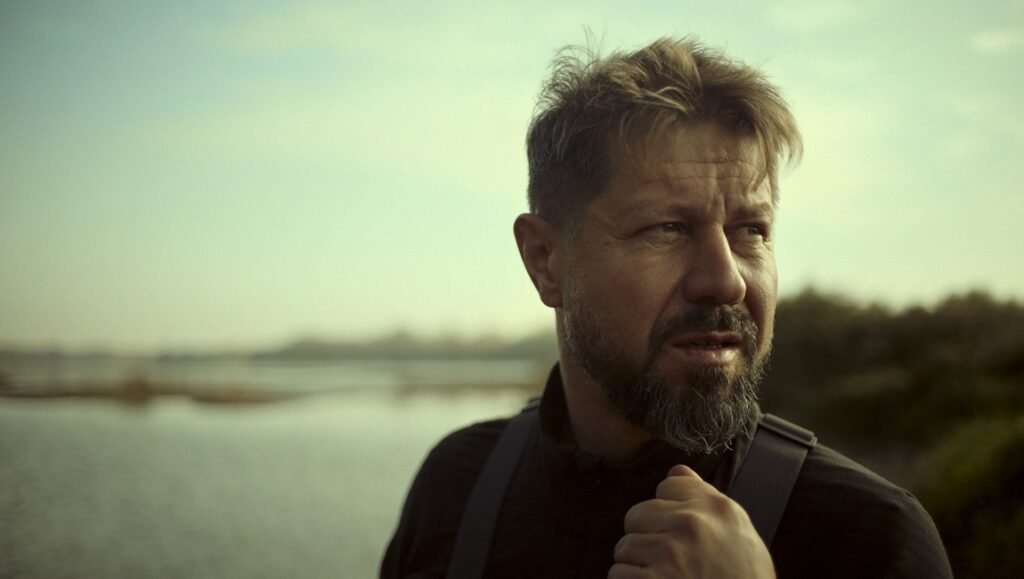 Grey-bearded man in a black top grips a strap and gazes across water toward a distant shoreline