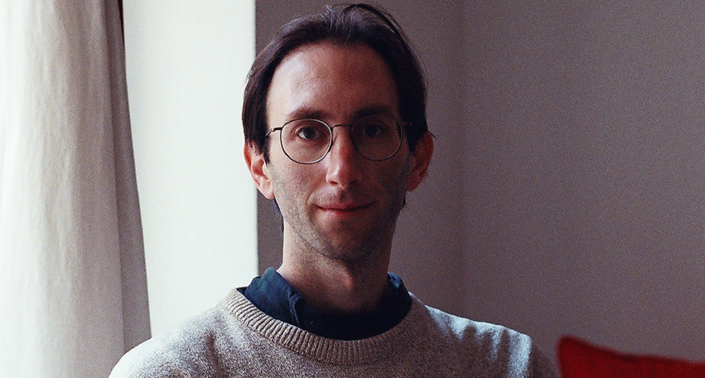 Man with round glasses and light stubble wearing a beige sweater and dark shirt, standing indoors near a white curtain.