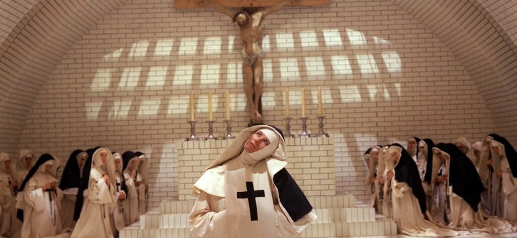 Nun in white habit with black cross raises her head before a crucifix as other nuns kneel in a vaulted brick chapel.