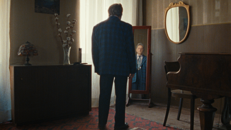 Older man in a dark plaid jacket stands with his back turned, facing an oval mirror in a dimly lit room.