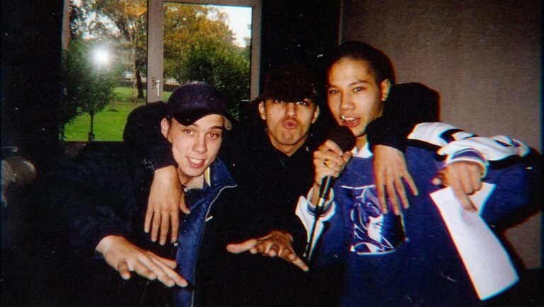 Three young men posing playfully indoors, one holding a microphone and another holding a paper, with a window showing trees in the background.