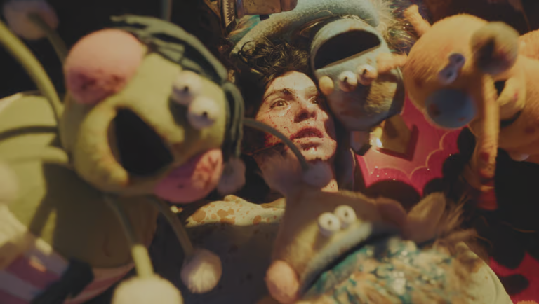 A woman lies surrounded by fuzzy puppets, one gripping her head in a dimly lit, surreal scene.