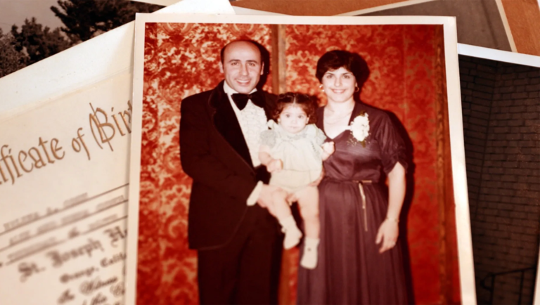 Vintage photo of a smiling man in a tuxedo and woman in a dark dress holding a baby against red floral wallpaper.
