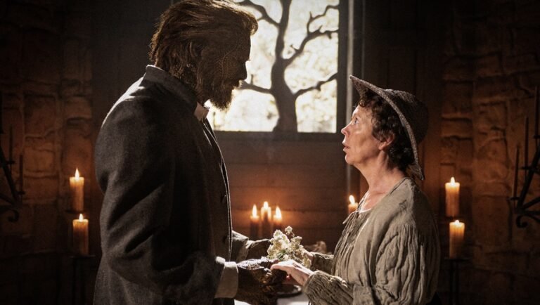 Beast-like man in a grey coat hands white flowers to a woman in a hat inside a candlelit stone room.