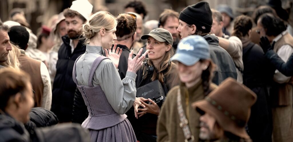 The Best Sex in Town: An Interview with Alex Huston Fischer & Eleanor Wilson on Wicker Costumed actress in a grey period gown gestures while talking with a crew member holding a monitor amid a crowded set.