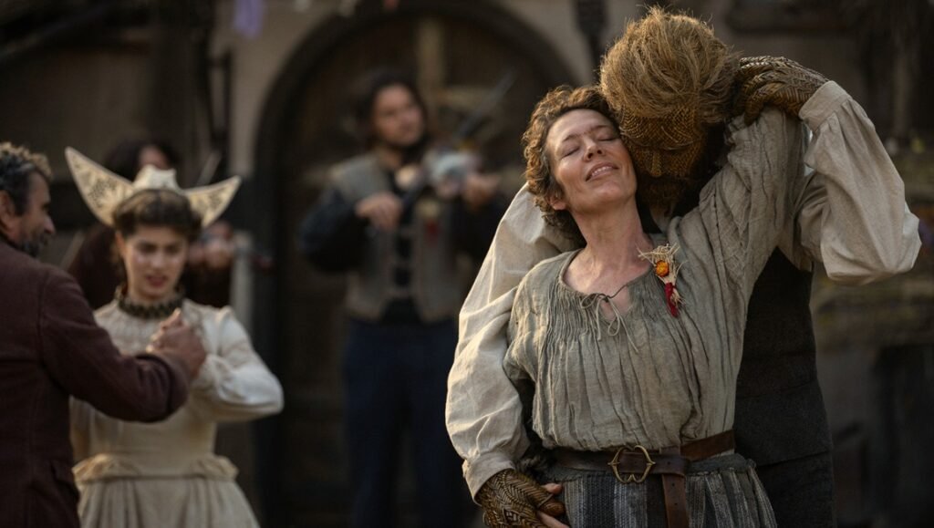 Smiling woman in a ruffled blouse is embraced from behind by a furry beast-man on a rustic street.