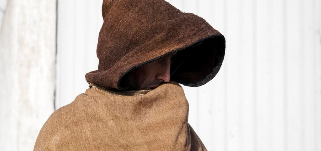 Figure in a brown hooded cloak turns away, face hidden, against a pale wooden wall.