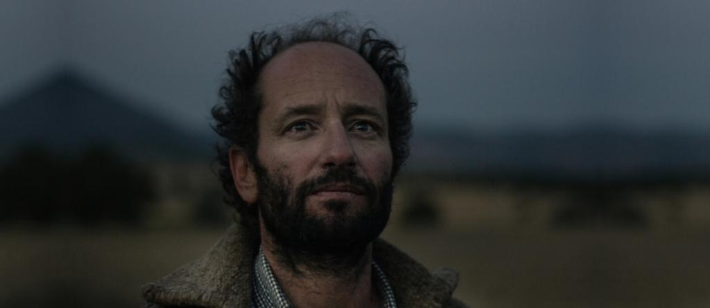 Bearded man with curly hair in a fur-collared coat gazes upward against a dim mountain landscape