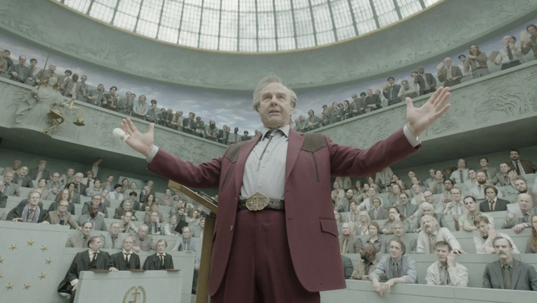 Man in a red suit with arms outstretched speaks before a large crowd in a domed hall.