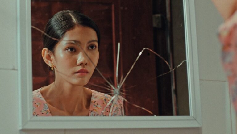Woman with slicked-back hair and gold earring gazes into a cracked mirror in dim warm light