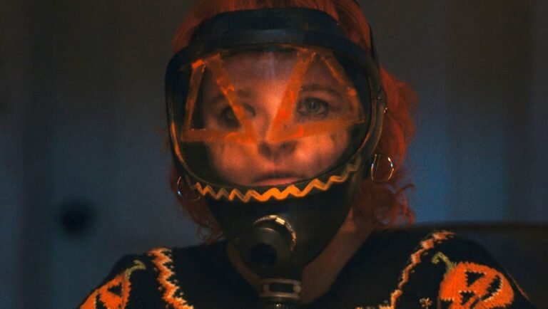 Person with orange hair wears a black helmet with an orange jack-o'-lantern face mask and a matching pumpkin-patterned sweater