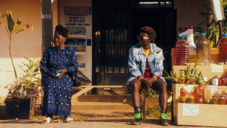 Woman in blue patterned dress sits beside youth in denim jacket outside a shop with stacked goods.