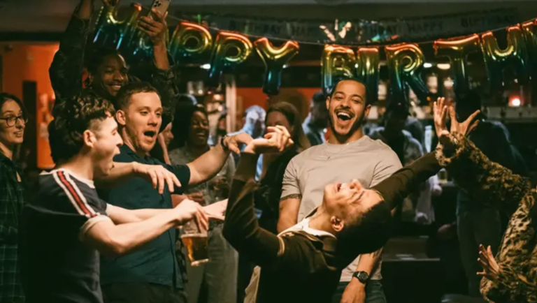 Man in a black suit leans back dancing as a cheering crowd raises a glass beneath a gold balloon banner