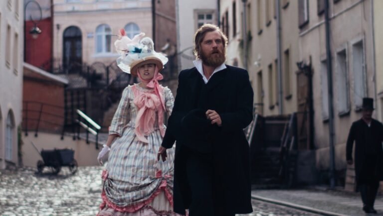 Bearded man in black coat walks beside woman in pink ruffled gown and flowered hat on cobblestone street