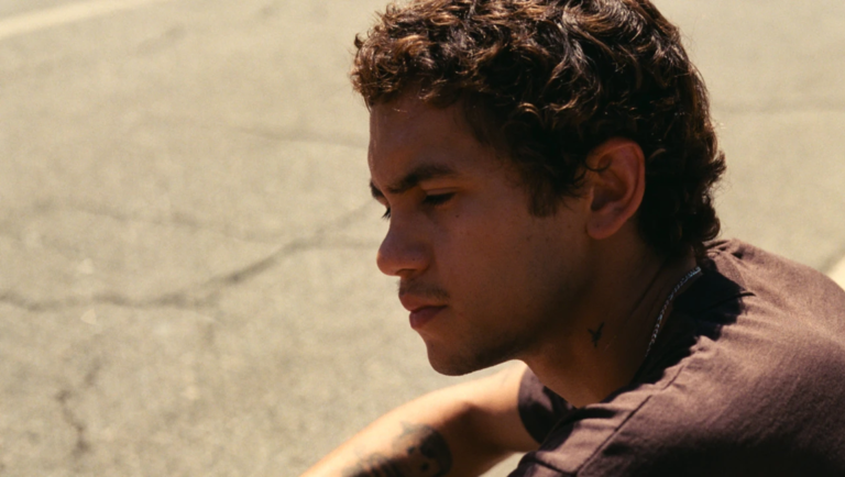 Close-up profile of a young man with curly brown hair looking down against a sunlit concrete wall.