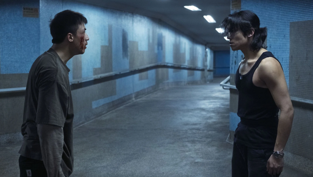 Two men face each other in a blue-lit corridor, one in a tank top and the other in a loose shirt.