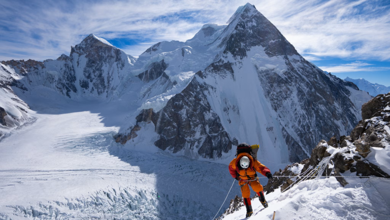 Mountaineer in orange suit ascends a snowy ridge with ropes, facing K2's rocky pyramid peak under blue sky.