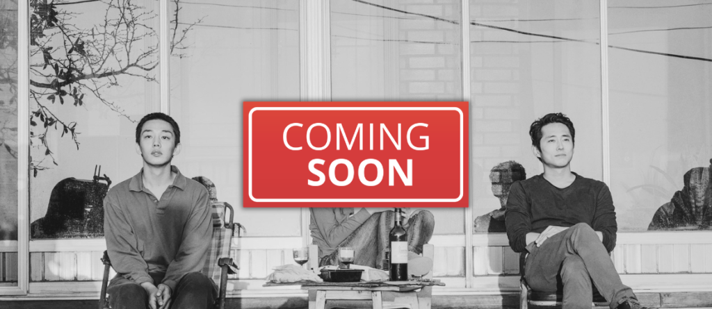 Two men sit apart on a porch outside a glass-walled building, a red 'COMING SOON' sign between them