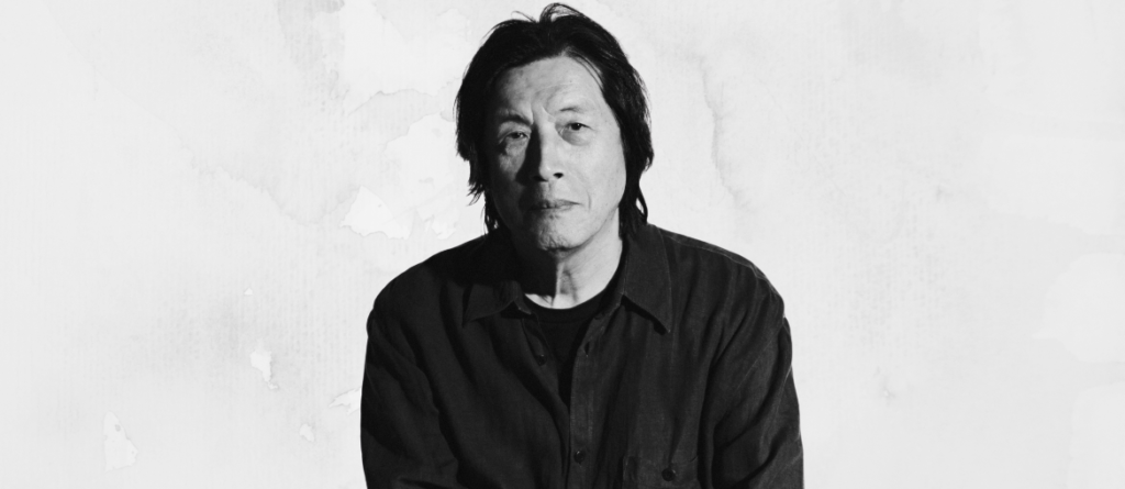 Black-and-white portrait of a man with shoulder-length dark hair in a buttoned shirt gazing at the camera