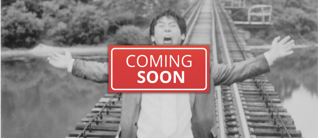 Person with arms outstretched on railway tracks, mouth open, behind a red 'COMING SOON' sign