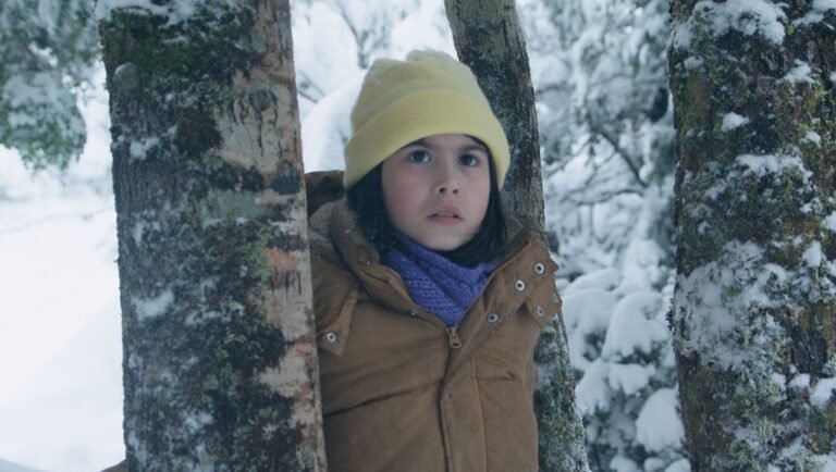 Young girl in yellow beanie and brown coat hiding between snow-covered tree trunks in a winter forest.