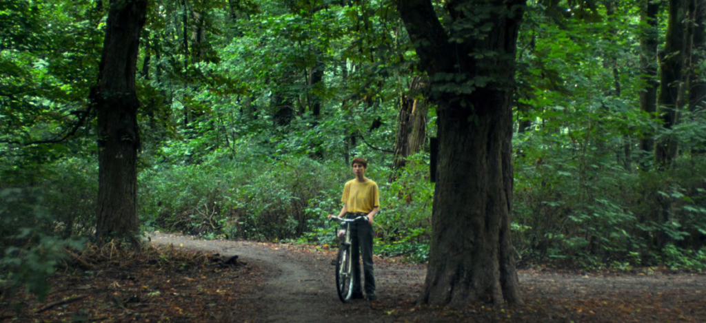 The Return of Language: My Wife Cries Within the Cinema of Angela Schanelec Man in a yellow shirt stands with a bicycle on a dirt path between large trees in a lush green forest.