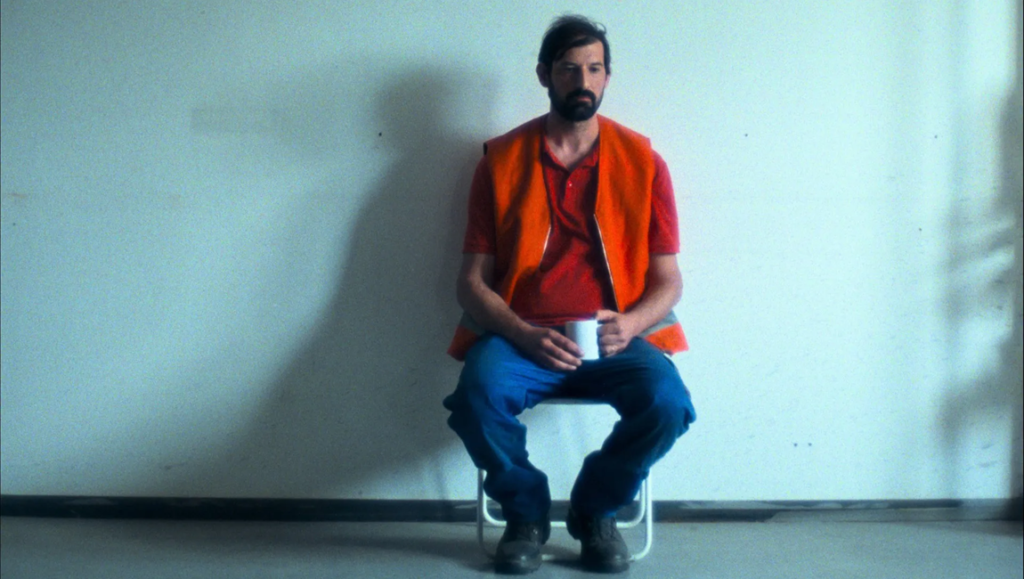 Bearded man in an orange vest and blue jeans sits holding a white mug against a pale wall.