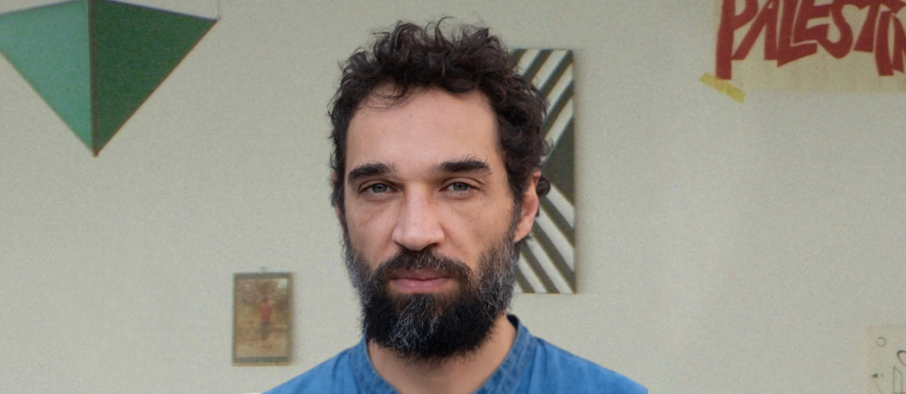 Bearded man with curly dark hair wearing a blue shirt, facing forward against a wall with geometric art.