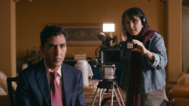 Man in suit and tie seated before a film camera operated by a woman wearing headphones in a warm-lit room