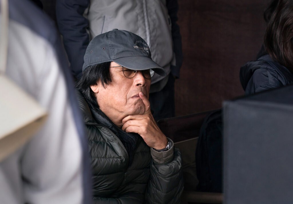 Bearded man in a navy cap and glasses rests his chin on his hand, wearing a black puffer jacket amid a crowd