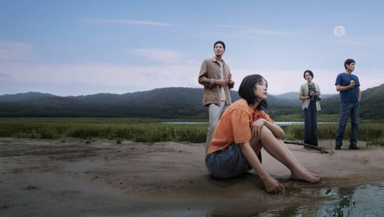 Woman in orange shirt sitting by water while three people stand along grassy shore beneath distant hills