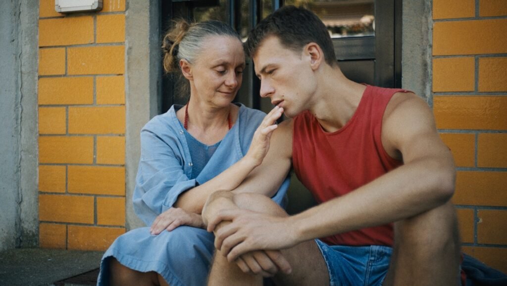 Woman in a blue dress touches the chin of a young man in a red tank top seated by a brick wall.