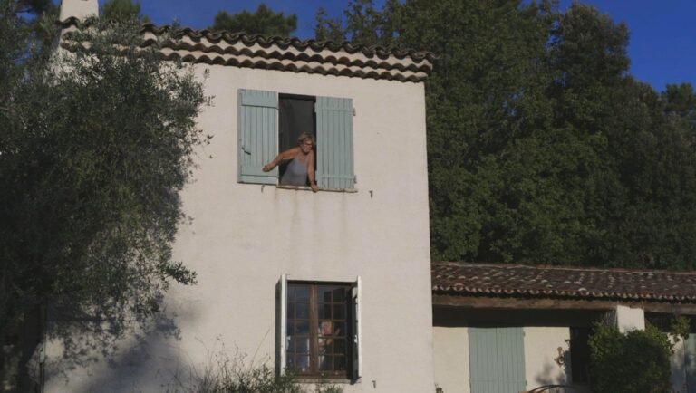 Woman leaning from a window with blue shutters in a white stucco house, surrounded by trees under a blue sky.