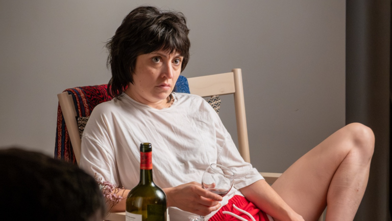 Woman with short dark hair in a white t-shirt sits holding a wine glass beside a green bottle, looking off-camera.
