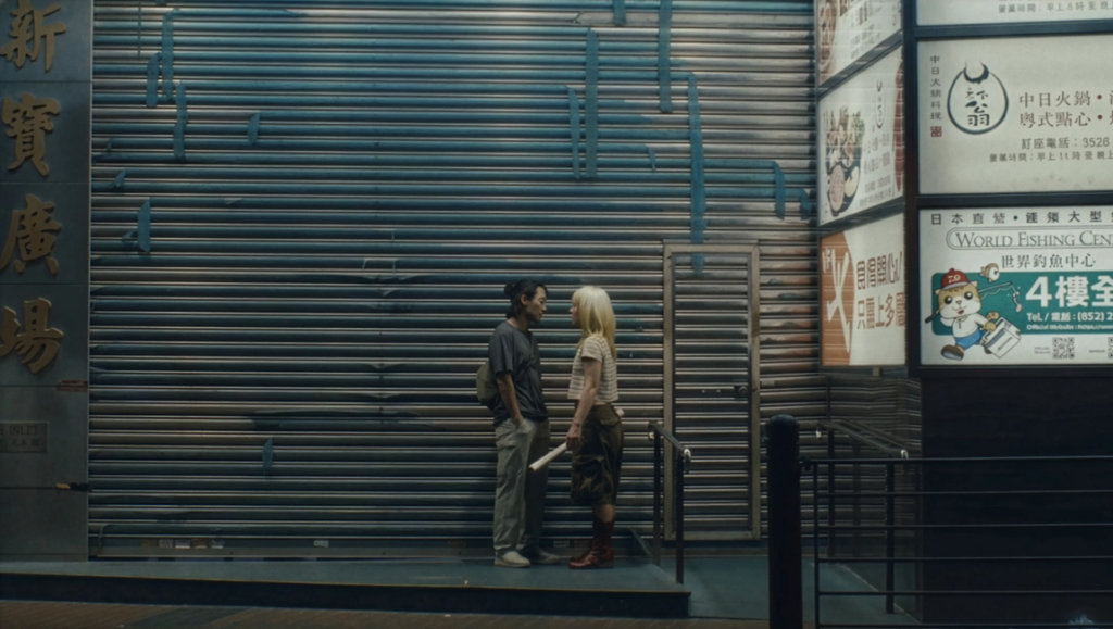 New/Next Film Festival 2026: zi, Sour Minnows, Here I’m Alive Man with a backpack and pink-haired woman holding a rolled paper face each other outside a shuttered storefront with Chinese signs